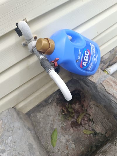 The image depicts an outdoor scene, likely in Ostrogorka, Russia, featuring a blue plastic jug of "ABC" brand cleaning liquid. The jug is positioned near a beige vinyl-sided wall and a concrete structure that appears to be a drain or a recessed area. A white pipe with a spigot is attached to the wall, and a red valve is visible on the pipe. The concrete area contains some debris, including leaves. The lighting suggests it is daytime. There are no people visible in the image.