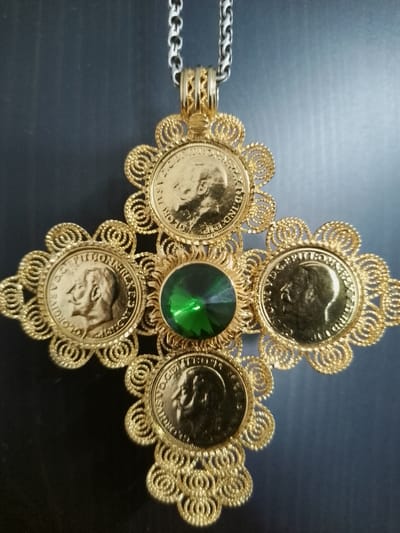 The image displays a close-up, overhead view of a gold-colored cross-shaped pendant with a large green faceted gemstone at its center. The pendant is suspended from a silver-colored chain. The cross is adorned with four gold-colored coins, each featuring a profile of a man's head. Intricate gold filigree work forms decorative elements around each coin and the central gemstone, resembling a flower or star shape. The background is a dark, textured surface, possibly a fabric or wood. The pendant is not located in the city of Nicorești, Romania.