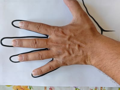 A close-up, top-down view shows a person's right hand and forearm resting on a white sheet of paper with a black outline of a hand drawn on it. The hand is positioned over the drawn outline, with the fingers slightly spread. The skin on the hand is light brown and shows visible veins and texture. The person's nails are clean and short. The background is a blurred white surface, and a floral pattern is visible at the bottom of the image. The scene is indoors, and no specific location within Crasnogorca, Moldova, can be determined.