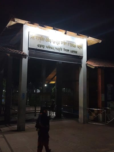 The image shows the entrance to a heritage site at night. The main structure features a wooden roof supported by a white pillar. A sign with golden text in Bengali is prominently displayed above the entrance archway, which reads "Shat Gombuj Masjidi O Bagerhat Sadar" (Sixty Dome Mosque and Bagerhat Sadar) and "Sanskriti Bicharak Sangrahalaya" (Cultural Exhibition Museum).

The foreground shows a person, possibly a security guard, walking away from the camera, wearing a dark uniform with orange accents on the pants. The ground is paved with bricks. Behind the entrance, there are metal fences and walkways, with visible stairs in the background. The overall environment suggests a public place that is illuminated by artificial lights at night. The inscription on the sign specifically identifies the location as the Shat Gombuj Masjidi (Sixty Dome Mosque) and Bagerhat Sadar, which is a UNESCO World Heritage site located in Bagerhat, Bangladesh, known for its historical Islamic architecture.