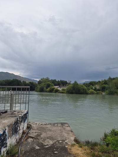 The image shows a view of the Karatal River in the city of Tekeli, Kazakhstan. The river flows from left to right across the frame. On the left side of the image, there is a concrete structure with a metal railing, which appears to be part of a dock or pier. The concrete is weathered and has graffiti on it. The river water is calm with small ripples. In the middle ground, there is a densely vegetated island in the river. On the far bank, there is a line of trees and some buildings. The background features rolling hills and mountains. The sky is overcast with dark clouds, suggesting impending rain. The overall impression is of a slightly overcast day with a natural landscape.