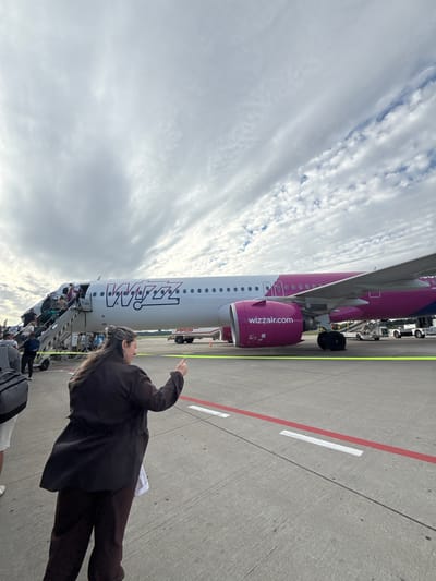 An exterior, low-angle, eye-level, medium shot shows a Wizz Air airplane at an airport in Künersberg, Germany. Passengers are boarding the plane via a set of stairs on the left side of the image. A woman in the foreground, facing away from the camera and to the right, gives a thumbs-up gesture. She is wearing a dark jacket and dark pants. The airplane is white with pink accents and the Wizz Air logo. The sky is cloudy. In the background, airport service vehicles are visible.