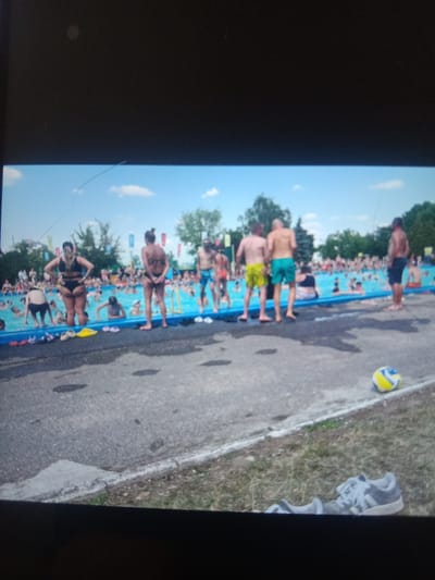 The image depicts a crowded outdoor swimming pool area, likely during warm weather. In the foreground, a paved surface transitions to a grassy area where a single white athletic shoe with grey laces and socks are placed. Further into the scene, a yellow, blue, and white volleyball rests on the grass. The main focus is a large, bright blue swimming pool filled with numerous people, many of whom are partially submerged in the water. Several individuals are visible standing at the edge of the pool, wearing swimwear. The background features trees and flags, suggesting a recreational park or water park setting. The lighting indicates daytime. The image does not contain any specific identifiable landmarks or features that would definitively place the location in the city of Kielce, Poland.