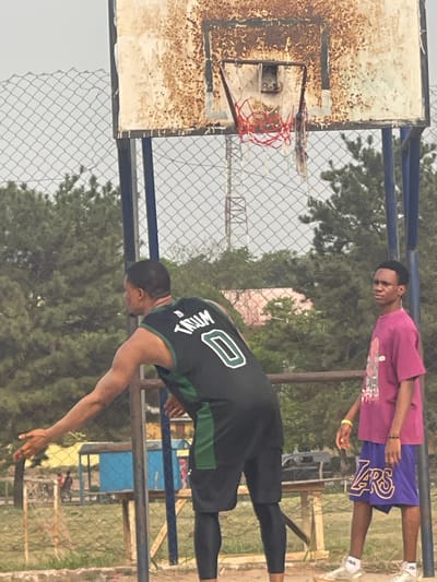 The image depicts an outdoor basketball court in Umunekwu, Nigeria, under an overcast sky. In the foreground, two adult males are visible. The individual on the left, viewed from the back, wears a dark green/black basketball jersey with "TRUM" and the number "0" in white, black shorts, and black compression tights. His right arm is extended downwards and slightly outwards, hand open. To the right, a second individual stands facing left, wearing a magenta t-shirt with a white graphic, purple shorts with "LAKERS" in gold lettering, and light-colored sneakers. A yellow wristband is present on his right wrist.
The basketball equipment includes a heavily rusted, peeling white backboard with a red rim and a partially torn red and white net. A dark blue support pole for the backboard is visible. Behind the court, a chain-link fence separates the playing area from the background. Visible through the fence are green trees, a distant metal lattice tower, a small structure with a blue base, and a light pink/orange building. The ground surface appears to be dirt or earth.
