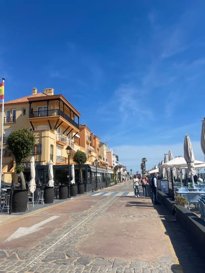 The image depicts a street scene in El Puerto de Santa María, Spain. On the left side of the frame are buildings with a yellow and orange facade, adorned with balconies and topped with red tiled roofs. Restaurant seating and umbrellas are visible along the street, with planters adding greenery.
On the right side of the image is a sidewalk and a street, marked with a white arrow. A few people are walking along the sidewalk. The sky is clear and blue. The overall impression is of a sunny day in a coastal city.