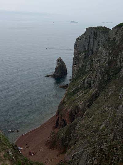 The image captures a coastal scene from Russkiy, Russia. A rugged cliff dominates the right side of the frame, extending downwards towards a sandy beach. A group of people is gathered on the beach. In the water, there are rock formations and boats. The sea stretches towards the horizon under a cloudy sky.