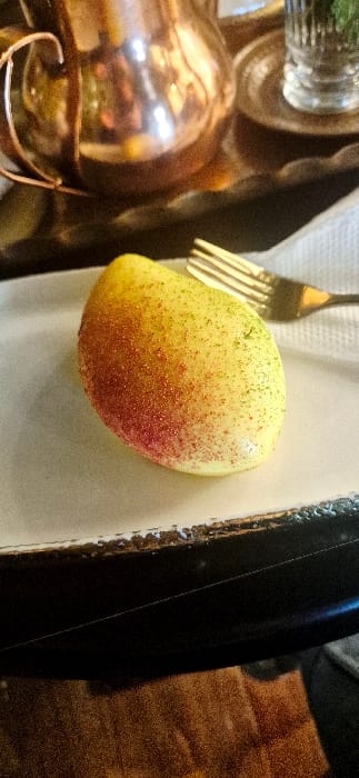 A ripe mango rests on a white plate alongside a golden fork. The mango's skin transitions from a light yellow to a vibrant red, speckled with tiny dots of color. A shiny copper-colored container is partially visible to the left. A white napkin partially obscures the container. A dark wooden surface is visible on the right.