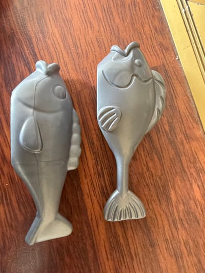 Two gray plastic fish figurines are displayed on a wooden surface. The figurine on the left is positioned vertically, while the figurine on the right is slightly tilted. The wooden surface exhibits a visible grain pattern. The background shows a golden-colored edge, possibly part of a table or frame. There is no indication of location in Ridabu, Norway, or any people present in the image.