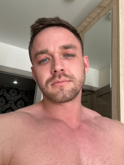 A close-up, slightly angled selfie of a fair-skinned man with blue eyes and a short beard is framed in the upper left corner by a light-colored wooden frame, possibly a mirror or picture frame. The man's upper chest and shoulder are visible, revealing reddened skin, possibly indicating sun exposure or irritation. The background includes a light-colored wall and a partially visible dark-colored door with a patterned surface, possibly wallpaper. The location is consistent with an indoor setting, potentially a bathroom or bedroom. There is no indication that the image was taken in Dobryanka, Russia.