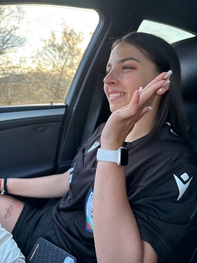 A young woman is seated inside a vehicle, possibly a car, as indicated by the window and interior trim visible. She has dark hair, light skin, and is smiling while raising her hand in a waving gesture. She is wearing a black sports top with a white logo and a white Apple Watch. A phone case with "KARL LAGERFELD" printed on it is visible. Part of a tattoo is visible on her leg. The background visible through the car window indicates a natural environment with trees and sunlight. The location is Gorna Rositsa, Bulgaria, as requested.