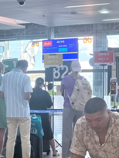 The image depicts a scene inside an airport terminal, likely in Mai Khao, Thailand, given the context of airline operations. In the foreground, a man wearing a floral-patterned shirt is seated and looking down. Behind him, a group of people are standing, appearing to be waiting. One person is wearing a light blue and white striped shirt and light-colored pants, another a black t-shirt and dark pants, and another a purple t-shirt and grey pants, with a light-colored backpack. A prominent digital display board shows flight information for AirAsia flight AK 819 to Kuala Lumpur, with the gate open and scheduled departure time of 08:40. There are also zone indicators for passengers, with "ZONE 2" and "ZONE 3" visible. The environment appears to be a boarding area, with glass partitions and typical airport amenities. The lighting suggests it is daytime.