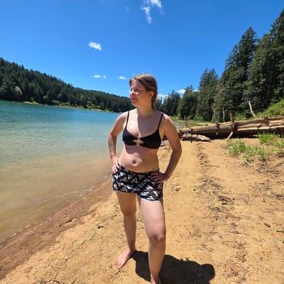 A woman stands on a sandy beach next to a body of water, with a forest of green trees in the background and a clear blue sky overhead. The woman is wearing a black bikini top and black and white patterned shorts. She has her hands on her hips and is looking to her left. The water is a light blue-green color, and the beach is made of light brown sand. There are some fallen trees and branches on the beach behind the woman. The image was taken on a sunny day. The location is a body of water in the city of Cherry Grove, United States.
