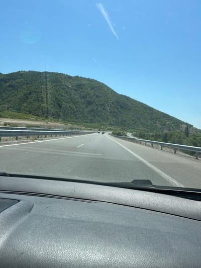 The image is a point-of-view shot from a moving vehicle on a highway. The road is asphalt with white lane markings and a metal guardrail on the right side. On the left, a lush green, forested hill rises steeply. The sky is clear blue with a faint contrail visible. The dashboard of the vehicle is in the foreground. The location is a highway near Jeleznitsa, Bulgaria.
