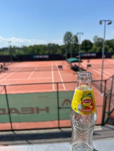 The image displays an outdoor tennis court facility located in Giebułtów, Poland, observed under bright daylight from an elevated vantage point, likely a spectator area. In the immediate foreground, an empty, clear 250ml glass bottle of Lipton Peach Flavour Fruity Ice Tea stands upright on a light grey concrete ledge. The bottle's yellow label is clearly visible, identifying the product.
The background, rendered out of focus, reveals multiple red clay tennis courts, each equipped with a white net. A dark chain-link fence separates the foreground from the playing areas. Attached to this fence is a green banner with a large, partially reversed "HEAD" logo. Tall metal light poles are positioned around the courts for illumination, and a green canopy structure is visible courtside on the right. In the far background, a dark banner or wall with white lettering is discernable, appearing to read "GIEBUŁTÓW PRO TENNIS," specifically identifying the tennis club in Giebułtów. Two indistinct human figures are faintly visible on the leftmost court near the net, suggesting activity. The scene is illuminated by a clear blue sky, casting shadows indicative of sunny conditions.