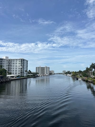 The media file presents a bright, sunny landscape view of a waterway in Delray Beach, United States of America. The image captures a canal or intracoastal scene with urban development and natural foliage along its banks under a blue sky.
The scene features a wide expanse of dark blue-grey water in the midground and foreground, showing distinct ripples and waves across its surface, reflecting the light and sky. On the left side of the waterway, several multi-story white buildings with numerous windows, likely condominiums or apartments, line the bank. A prominent, modern building stands in the left foreground, while other similar structures recede into the distance. Docks and a few boats are visible along this bank, some partially obscured by the buildings.
The right bank of the waterway is characterized by lush, vibrant green palm trees and other tropical vegetation. Amidst this greenery, some residential structures or houses can be faintly seen. A few boats are also docked along this tree-lined shore.
The sky, occupying the upper portion of the image, is a clear, bright blue with scattered white, wispy clouds that appear streaky and dispersed. The overall weather appears sunny and pleasant.
No specific activity or event is prominently taking place; the scene depicts a tranquil, static view of a waterfront area. The bright illumination suggests it is daytime, likely late morning or early afternoon. The architecture, palm trees, and waterway setting are consistent with coastal Florida environments like Delray Beach. There is no visible text in the image.