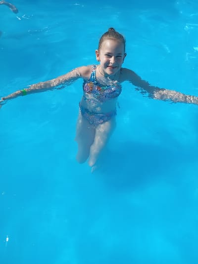 A young girl with her hair in a bun is floating on her back in a bright blue swimming pool. She is wearing a colorful swimsuit with a floral pattern. Her arms are extended to the sides, and she is smiling at the camera. The water is clear and illuminated by sunlight, suggesting it is daytime and the weather is pleasant. Another person's arm, wearing a green wristband, is visible in the upper left corner of the image, also in the water. The scene appears to be a recreational setting, possibly a hotel pool or a public swimming area.