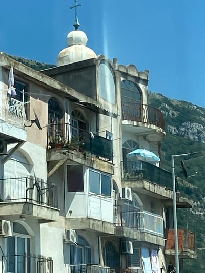 The image shows a multi-story building with a domed structure and a cross on top, located in Mišići, Montenegro. The building appears to be made of concrete and has several balconies with metal railings. Some balconies have plants, and one has a blue umbrella. There are windows and air conditioning units visible on the building's facade. The building is situated on a hillside, with a green, mountainous landscape in the background. The sky is clear and blue. There are no people visible in the image.