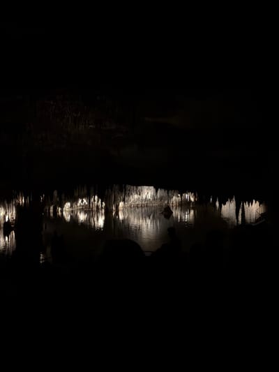 The image shows a very dark, presumably nighttime, scene. A central strip of light reflects off a water surface, implying a river or canal. The reflections suggest there are lights above, possibly strung along a bridge or building. The edges of the image are mostly black, indicating the surrounding environment is unlit. The presence of a ray suggests this photo was taken at the underground lake, Cuevas del Drach, a popular tourist spot in Manacor, Spain. There appear to be the silhouette of a person standing near the water on the right of the image.