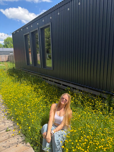 The image captures a young woman sitting in a lush field of vibrant yellow wildflowers under a bright, clear sky. She has long, straight blonde hair and is wearing a white crop top, light blue jeans, and sunglasses. She is positioned towards the right side of the frame, with her arms resting on her knees, and she looks towards the viewer with a relaxed, slightly smiling expression.

To the left of the woman, a modern building with dark grey corrugated metal siding is prominently featured. It has several horizontal windows that reflect the blue sky, scattered white clouds, and green foliage. A string of decorative light bulbs hangs along the eave of the building. A wooden deck or walkway is visible in the foreground, at the bottom right.

The scene is illuminated by bright sunlight, indicated by the crisp shadows cast. The weather appears pleasant and sunny. The overall impression is one of leisure and enjoyment of the outdoors in a natural setting, potentially in Vozhoiskii, Russia. There is no visible text in the image.