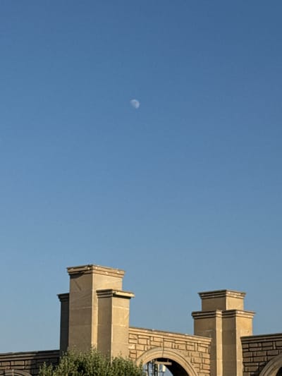 This media file shows a low-angle shot of a large, light-colored stone monument against a clear blue sky. The monument features geometric architectural elements, including stepped ledges and decorative brickwork patterns. On the right side of the frame, a section of lush green foliage is visible, suggesting the monument is situated outdoors, possibly in a park or garden. In the upper left quadrant of the sky, a small, crescent-shaped moon is faintly visible. The clear sky and the brightness of the monument suggest it is daytime, likely late morning or early afternoon given the shadows. The overall impression is one of grandeur and permanence. The monument's style, along with the foliage and clear sky, could suggest a location in a warm climate. No text is visible in the image.