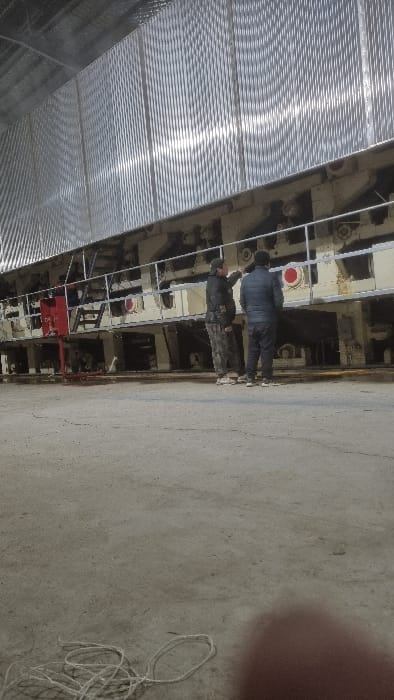Two individuals are standing near industrial machinery inside a warehouse. One wears a dark jacket, while the other is in camouflage. The machinery appears to be attached to the warehouse's side wall. The warehouse features a corrugated metal roof. The floor is concrete, marked with a crack. A blurred object is present in the upper right corner of the image, alongside a tangled white cord on the floor.