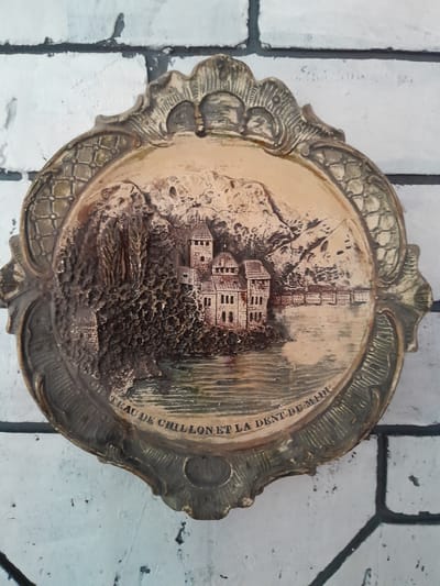 The image shows a decorative plate mounted on a white brick wall. The plate is oval-shaped with an ornate, embossed border. The central image on the plate depicts a castle situated on a rocky landscape next to a body of water, with mountains in the background. The text "CHÂTEAU DE CHILLON ET LA DENT DU MIDI" is inscribed along the bottom edge of the plate, indicating the scene depicted is Château de Chillon and the Dent du Midi mountain in Switzerland. The plate is the sole subject of the image and it is not related to the city of Cimișlia, Moldova.