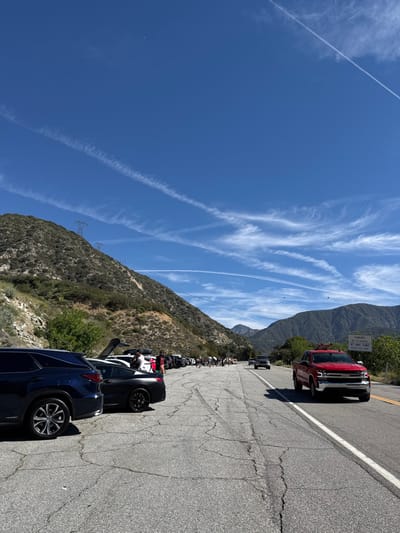 The image depicts a sunny, high-altitude roadside scene in the San Gabriel Mountains, consistent with Angeles Crest Highway (SR 2) within or near La Cañada Flintridge, California. The sky is deep blue, featuring multiple distinct white contrails and wispy cirrus clouds. Mountains covered in dry, light-green brush and sparse trees rise steeply on both sides of the cracked asphalt two-lane road, which shows visible tire marks. Power lines with transmission towers are present on the left mountainside.
In the foreground on the left, a dark blue SUV, appearing to be a Lexus RX, is parked with its rear towards the viewer. Immediately adjacent, a dark grey two-door coupe, possibly a BMW 8 Series, is also parked. A long line of approximately 20-30 additional parked vehicles, including sedans, SUVs, and coupes, extends into the distance along the wide shoulder on the left side of the road. Many of these vehicles have open trunks or tailgates. Scattered among these parked vehicles are approximately 20-30 individuals, some partially obscured, standing and congregating.
On the right lane of the road, a red Chevrolet Silverado pickup truck with a chrome grille is driving towards the camera. Further down the road, several other vehicles are visible, including a dark SUV/truck moving away from the viewer. A white rectangular sign with black text, reading "CHAINS MAY BE REQUIRED AT ANY TIME," is posted on the right side of the road in the mid-ground. The scene portrays a large gathering of people and vehicles in a mountain pass environment.