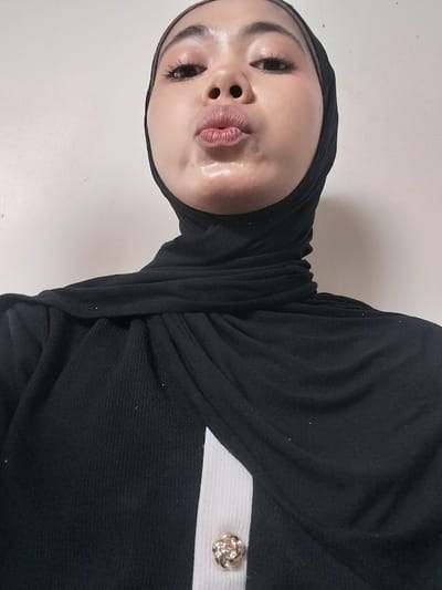 A close-up, chest-up portrait shows a young person wearing a black hijab and a black garment with a white accent and a metallic button. The person's face is tilted upwards, and their lips are pursed as if blowing a kiss. Their makeup is visible, with emphasis on the eyes and lips. The background is a plain, light-colored wall. The image was taken indoors. There is no information available about the location in Jeppee, Indonesia.