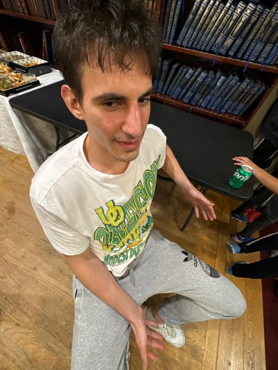 A young adult male, appearing to be in his twenties, with dark, slightly disheveled hair and a light sheen on his face, is captured in a high-angle shot, looking downwards and to his right with a subtle facial expression. He is wearing a white short-sleeved t-shirt with a green and yellow "Oregon Ducks" logo, gray Adidas sweatpants, and white athletic shoes. His hands are open and positioned low, palms slightly upward. The subject is situated on a light-colored wooden floor.
In the background, to the subject's right, a dark, rectangular table holds a green Sprite soda can. A child's hand is visible near the can. Below this table, the lower legs and feet of multiple individuals, likely children, wearing various colored sneakers, are partially visible. To the subject's left, a table draped with a white tablecloth displays several white rectangular trays containing food items, including what appear to be appetizers with green garnishes. Behind these elements, a dark wooden bookshelf extends across the upper right background, filled with numerous uniformly bound blue books featuring gold lettering on their spines. This environment is consistent with a library or study hall within a religious institution in Bergenfield, United States.