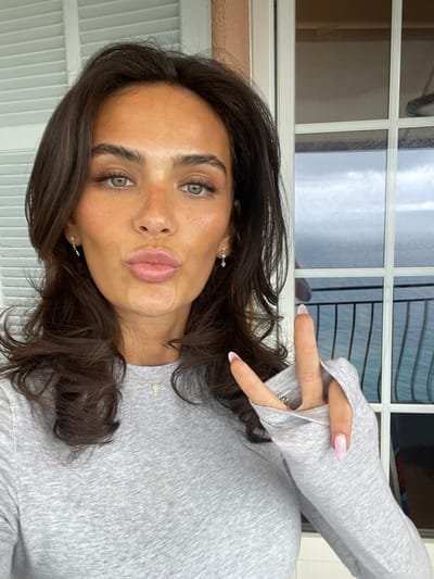 The image shows a woman with long, dark brown hair, wearing a gray long-sleeved shirt, making a peace sign with her right hand. Her left hand is not visible, but she appears to be wearing small earrings. She is standing in front of a window with a view of the sea, likely taken on a balcony in Roquebrune-Cap-Martin, France. She is making a kissy face. The weather is cloudy, and the sea is visible in the distance. The woman appears to be in her twenties, with a youthful and healthy appearance. The overall impression is of a carefree and cheerful woman enjoying the view from her balcony.  The balcony is supported by a white balustrade. The image is a selfie, likely taken with a smartphone.