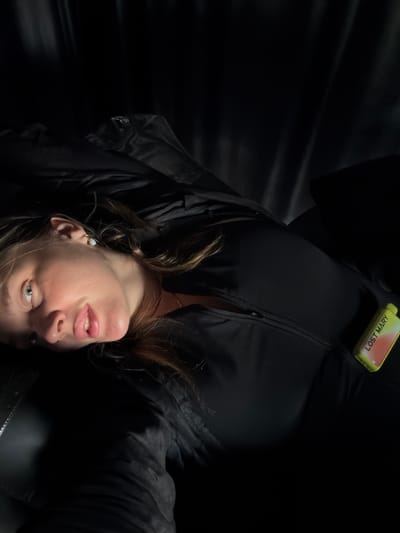 A woman is lying on her back on a dark surface, possibly a couch or bed, with her head turned towards the camera. She has light brown hair, light blue eyes, and full lips. She is wearing a black top and a jacket or coat, also black. A vape device with the label "LOST MARY" is visible tucked into the front of her clothing. The background is dark and indistinct, with fabric folds visible, suggesting an indoor setting. The lighting is dim, with a spotlight illuminating her face and upper body. There are no visible indications of the specific location in Укромновское сельское поселение, Ukraine.