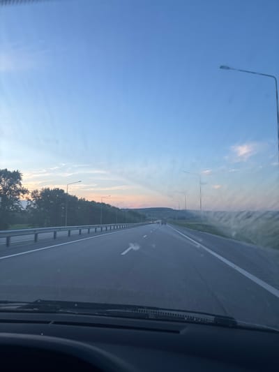 The image captures a view from the driver's perspective through a car windshield, looking out onto a multi-lane highway. The scene appears to be set in the early evening, as indicated by the soft light and the pastel colors in the sky, transitioning from blue overhead to hues of pink and orange near the horizon. Tall trees line the left side of the highway, casting shadows. Several streetlights are visible along the road. Distant hills are visible in the background. The road itself is paved and marked with white lines, suggesting it is a well-maintained thoroughfare. A few vehicles are seen in the distance on the highway. The overall environment suggests a rural or semi-rural setting, possibly on the outskirts of a town or city. The presence of a highway indicates a location where transportation and travel are prevalent. The image is captured at an angle from inside a vehicle, with the dashboard and windshield wipers partially visible in the foreground, adding to the sense of being in transit. The location is specified as Novye Karamaly, Russia.