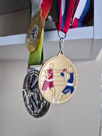 A close-up shot shows three medals hanging from a white shelf. The uppermost medal is circular and gold, depicting two boxers. One boxer, in a blue uniform, is standing, while the other, in a red uniform, is on the ground. The medal is attached to a ribbon with red, white, and blue stripes. Below this is a medal with a green ribbon and a gold edge. This medal is also circular and appears to have a circular emblem in the center with a bird-like figure and stars. The third medal, partially visible, is larger and appears to be made of a dark, reflective metal with a faceted design. The medals are hanging indoors, against a light-colored, out-of-focus background. The lighting suggests it could be daytime, but there are no specific cues to determine the time of day or weather. No text is visible on the medals. The location is identified as Lenina, Russia.