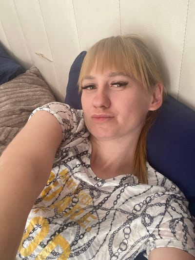 A blonde-haired woman is reclining in a bedroom, resting her head on a dark blue pillow. She is wearing a white t-shirt with a black and gold chain pattern and the word "SUPER" visible. The room has white paneled walls. A taupe-colored pillow is visible in the bottom left corner. The location is Vasyurinskaya, Russia. No specific actions or interactions are visible.