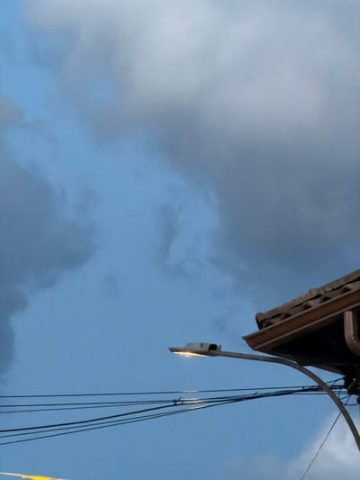The image captures an outdoor scene dominated by a cloudy, deep blue sky, likely during twilight. In the foreground, a section of a brown-tiled roof with a gutter is visible on the right side. A street lamp with its light on extends from the right, curving towards the center of the frame. Below the lamp, several black wires are strung horizontally across the sky. The overall impression is one of an overcast evening in an urban or suburban environment. The location within the city of Arcahueja, Spain, is not explicitly depicted through any identifiable landmarks.