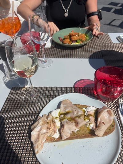 The image shows a table setting at a restaurant in Tignes, France, at the coordinates 45.454 latitude and 6.898 longitude. The table is set for two people, with two glasses of wine, one glass of orange juice, and two place settings. The table is covered in a white tablecloth with a brown placemat. A plate of food with an open faced sandwich with mortadella, pesto and burrata, is placed on the table, and to the side is an empty green plate. A red glass sits on the table. Two people are visible in the background. They are seated at the table, one is wearing a black top and the other is wearing a white shirt. One of them is holding a glass of wine and the other is holding a glass of orange juice. The person in the black top has a necklace and a bracelet on their wrist. The restaurant has a white floor with a light brown checkered tile pattern. There are other tables in the background, also with white tablecloths. It is a sunny day and the light is bright.