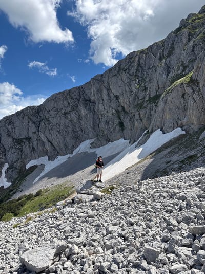 In Pašina voda, Montenegro, a person stands on a rocky slope with a large, grey, mountainous incline dominating the background. Patches of white snow are visible against the rock face. The sky above is blue with scattered white clouds. The person, wearing a hat, black shirt, and light-colored shorts, is positioned mid-frame, facing towards the camera. The foreground consists of a dense scattering of grey rocks. The overall scene depicts a mountainous outdoor environment.