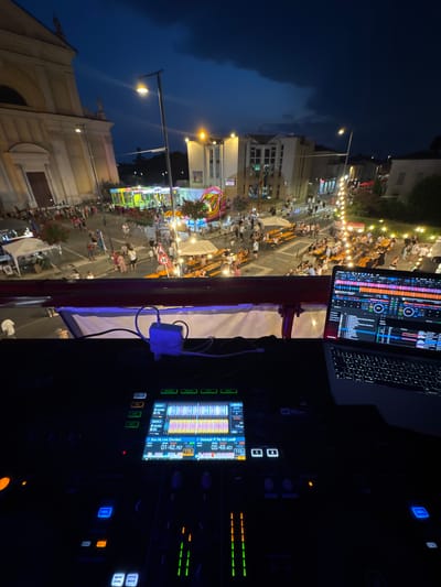 This is a first-person perspective, eye-level shot of a DJ setup at an outdoor evening event in the city of Calcio, Italy. The DJ booth is in the foreground, showing a laptop displaying DJ software with waveforms, and a DJ mixer with illuminated buttons and screens. Cables and power adapters are visible connected to the equipment.
In the middle ground, the scene opens up to a lively town square or street at dusk. A crowd of people is gathered, many seated at long wooden tables, suggesting a communal dining or gathering area, possibly related to a local festival or market. String lights are hung overhead, creating a festive atmosphere. Buildings, some with illuminated windows, line the street, and a large church or public building with a facade is visible on the left. In the background, a large inflatable structure, likely a bouncy castle, suggests entertainment for younger attendees. The sky is a deep blue, indicating nighttime. The overall environment is a public space, bustling with activity under artificial lighting as darkness falls.