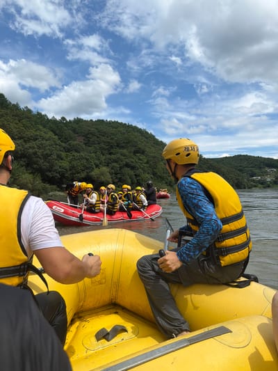 The image depicts a group of people participating in a white-water rafting activity on the Geumgang River in Seo-myeon, South Korea. The perspective is from within a yellow inflatable raft, looking towards another red inflatable raft carrying several individuals. Both rafts are equipped with paddles, and the participants are wearing yellow safety helmets and bright yellow life vests. The people in the red raft are actively paddling, suggesting movement through the water. The environment is characterized by a wide, calm river with a backdrop of lush green, tree-covered hills and a partly cloudy sky. The overall scene conveys a sense of outdoor recreation and adventure.