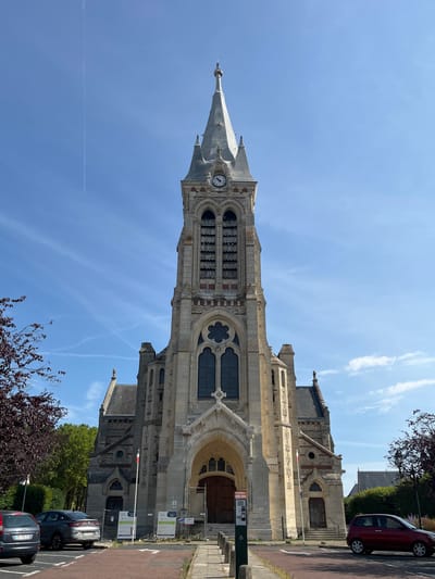 The image shows the facade of the Saint-Lubin church in Rambouillet, France, on a clear day with a blue sky and some wispy clouds. The church is constructed of light-colored stone and features a tall, ornate bell tower with a spire and a clock face. The main entrance is a large arched doorway with wooden doors. To the left and right of the main entrance are smaller arched doorways. There are trees with dark red leaves on the left side of the image and trees with green leaves on the right. Several cars are parked in a paved area in front of the church, and some temporary construction barriers and signs are visible. A red car is driving away from the church on the right side of the image.