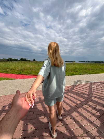A first-person perspective shot shows a person's hand holding another person's hand as they walk along a brick-paved path. The person whose hand is being held is facing away from the camera and is wearing a light blue t-shirt and shorts set with patterned slip-on shoes. Their blonde hair is visible. The path is curved and casts a shadow pattern on the ground. Beyond the path, there is a strip of red gravel followed by a field of tall green grass and reeds. In the distance, there is a body of water, trees, and a cloudy sky. The location is specified as Breytovo, Russia.