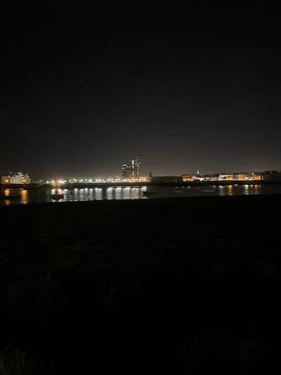 A panoramic nighttime photograph captures the waterfront cityscape of Rochester, United Kingdom. The immediate foreground is dominated by a dark, indistinct landmass or low-lying vegetation. A body of water, consistent with a river or estuary, spans the midground, reflecting numerous artificial lights from the opposite shore as elongated, shimmering vertical streaks. Several small, dark-hulled watercraft are visible on the water's surface. The background presents the city's illuminated skyline, featuring a continuous line of structures. A prominent cluster of modern, multi-story buildings with numerous lit windows rises centrally, flanked by other buildings of varying heights and architectural styles, all contributing to the ambient light reflected on the water. The sky above is dark, with only a few faint, scattered points of light discernible. No individuals are visible in the scene.
