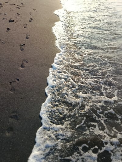 The image shows a black sand beach in Arjasa, Indonesia (latitude: -7.71, longitude: 114.117). The right side of the image shows a foamy wave receding back into the water. The left side of the image shows a series of footprints in the black sand. The footprints lead toward the receding wave. The sun is shining on the scene.