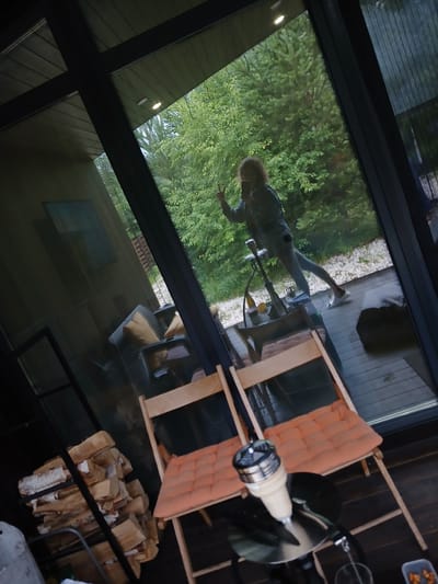 This media file captures a reflection in a large glass window, likely from the interior of a modern house or cabin. The reflection shows a person standing outside, outdoors, with lush green trees and foliage behind them, suggesting a natural setting. The person appears to be in casual attire, with one leg extended and arms outstretched, possibly in a playful or relaxed pose. Their hair is light-colored and appears to be curly. The scene outside is bright, indicating it is daytime, and the weather seems pleasant, with no visible rain or harsh sunlight.

Inside the house, reflected in the glass, are various objects, including a wooden chair with an orange cushion, stacks of firewood, and what appears to be a metal stand with a decorative element that resembles a spinning fan or a propeller. There's also a glimpse of furniture and interior structures, hinting at a contemporary living space with natural wood finishes. The overall mood is one of leisure and connection with nature. The image is vertically oriented, with the reflection of the person appearing somewhat distorted due to the angle and the nature of the glass. The context provided suggests this scene is in Verkhneye Dubrovo, Russia.