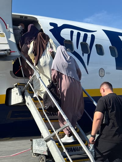 In the city of Kruja, Albania, on a sunny day, a group of people are boarding a Ryanair airplane via a portable staircase. The plane's fuselage is white with a large dark blue graphic. Several individuals, predominantly women wearing hijabs in various colors including purple, brown, and grey, are ascending the stairs. One woman in a beige dress is also visible. A man in a black t-shirt and black pants is descending the stairs, holding a bottle of water. The staircase is made of metal with yellow safety markings on the steps, and the Ryanair logo is visible on the steps. A small utility cart with the number 107 is positioned at the bottom of the stairs. The ground appears to be tarmac.