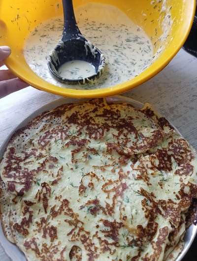 A close-up shot captures a portion of the cooking process for a batch of savory pancakes or crepes, likely in a home kitchen setting in Нижнетроицкий, Russia. In the upper portion of the frame, a bright yellow bowl overflows with a creamy, pale batter speckled with finely chopped green herbs, possibly dill. A black ladle, partially submerged in the batter, is being lifted out, showcasing a dollop of the mixture clinging to its edge. The hand holding the bowl is visible on the left, with fingers gently gripping the rim.
Below the bowl, a stack of golden-brown pancakes or crepes fills the lower half of the image. They have a slightly irregular shape and a characteristic lacy appearance due to the cooking process, with areas of crisp, browned edges and lighter, softer centers. Green herb flecks are also visible within the cooked batter, indicating they were incorporated into the pancake mixture. The pancakes are arranged on a white plate, which is partially visible. The background consists of a light-colored wooden surface, suggesting a table. The overall lighting suggests daytime, with no specific weather conditions discernible. The scene evokes a sense of domestic cooking and preparation of food. No visible text is present in the image.