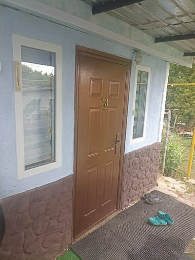 The image shows the exterior of a building, possibly a small house or a shed, with a brown front door and two windows. The building's facade is painted a light blue and the lower portion is decorated with a brown stone-like pattern. A metal roof overhang covers the entrance area. On the doormat, there are two pairs of sandals: one black pair and one pair of blue clogs. To the left of the door, a thermometer is attached to the wall. The background shows greenery and trees, suggesting an outdoor setting. The door is numbered "13".