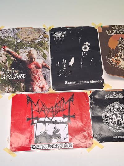 The image displays a collection of posters on a white wall, likely in a casual setting like a bedroom or studio. The posters are attached with yellow tape and pushpins. They are predominantly black and white, with one poster featuring a vibrant red background.
The posters feature a variety of graphic imagery and text, suggesting they are related to music or art. One poster shows a black and white illustration with a skull and crossbones motif and what appears to be text in an unfamiliar script. Another black poster has a stark white image of a face with a circular eye. Below this, a poster with the text "Transilvanian Hunger" in an ornate font is visible. A bright red poster dominates the right side, with a black, intricate logo and text that reads "DEATHCRUSH" in a gothic font. In the bottom left, a poster depicts a person lying on the ground, covered in what appears to be blood or red paint, with the word "lifelover" written in white script. The setting appears to be outdoors, with grass and rocks visible in the background of this poster. The overall aesthetic of the posters suggests a dark or heavy genre of music or art. There are no clear indications of time of day, weather, or specific emotions conveyed by the people depicted, beyond the general tone of the artwork.
