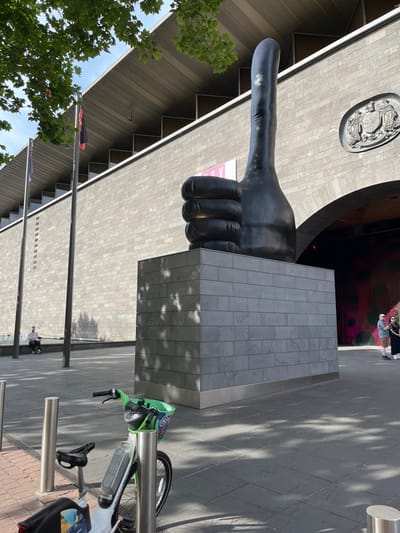 This outdoor photograph, rotated 90 degrees counter-clockwise, depicts a prominent public sculpture in an urban setting, identified as Southbank, Australia.
The central subject is a colossal, dark grey sculpture of a "thumbs-up" hand, resting on a sturdy, tiled plinth made of light grey, possibly metallic, material. This sculpture, known as "Monument to the Great Australian Thumbs Up" by Tony Clark, is a distinctive landmark.
In the background, a modern building with a textured, light grey facade serves as the backdrop. Above and to the left of the sculpture, a circular relief carving is affixed to the building's wall. A large, dark, curved archway, possibly an entrance, is visible to the right of the sculpture. Faintly visible on the building behind the sculpture is a pinkish sign with the letter "V", possibly part of the "ACCA" (Australian Centre for Contemporary Art) branding.
The setting appears to be a paved public square or walkway, with various shades of grey paving stones. Several tall, slender metal poles, potentially flagpoles, are visible to the left. On the right side of the image, a shared bicycle or e-scooter, with noticeable green accents, is parked in a metal bike rack. The bicycle has some branding, but no clear text is legible.
Sparse green foliage from trees is visible on the far left, suggesting the presence of greenery in the area. Two individuals are seen near the dark archway in the background, appearing to be walking or standing. Another person is visible further back, near the bike rack.
The time of day appears to be sunny daytime, with clear skies, indicated by bright lighting and distinct shadows cast by trees and objects onto the pavement and the sculpture's plinth. The overall scene suggests a vibrant, modern urban environment.