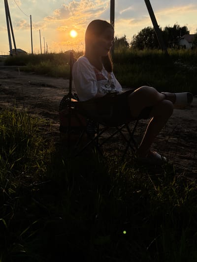 A young person is sitting in a folding chair, outdoors, with their legs crossed. The sun is setting, casting a warm glow over the scene. The person is wearing a white t-shirt with a graphic print. They are positioned in a way that suggests relaxation and contemplation. The background shows a natural landscape with grass and trees, indicative of a rural or semi-rural setting. Power lines and poles are visible in the distance, suggesting proximity to infrastructure. The overall mood is serene and peaceful, enhanced by the golden hour lighting. The image is taken from a low angle, focusing on the person and the surrounding environment. The location context is identified as Пропан Снт, Russia.